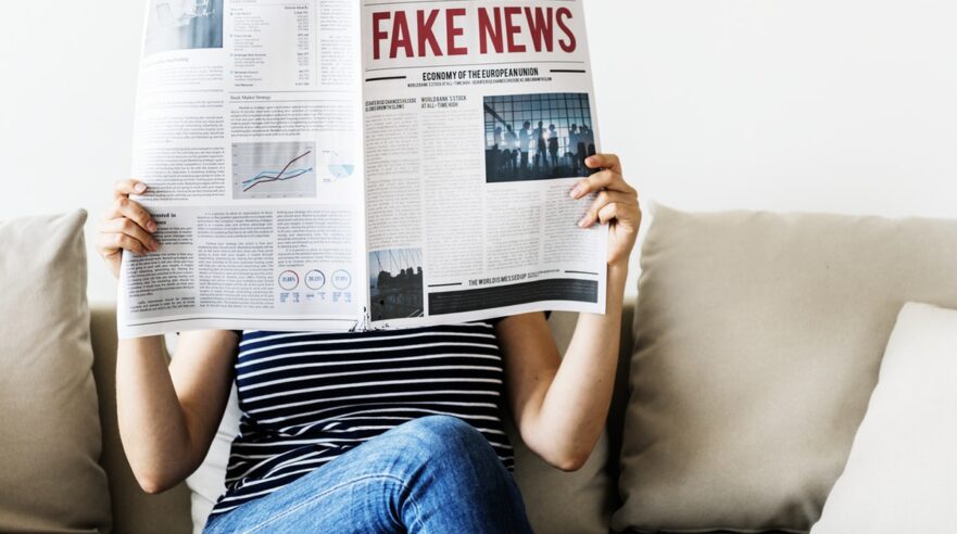 Iemand op de bank, benen over elkaar gekruist. Leest een krant met grote rode letters met Fake News erop.