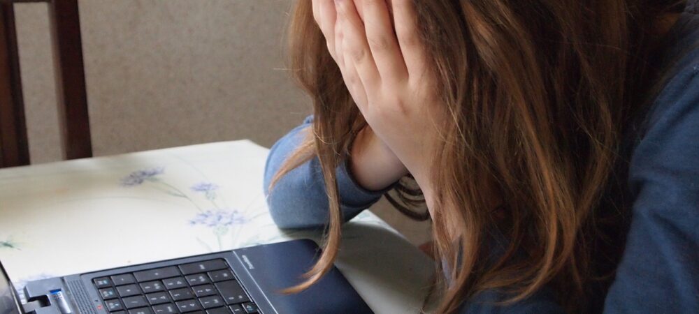 Mens met lang blond haar met handen voor gezicht achter laptop.